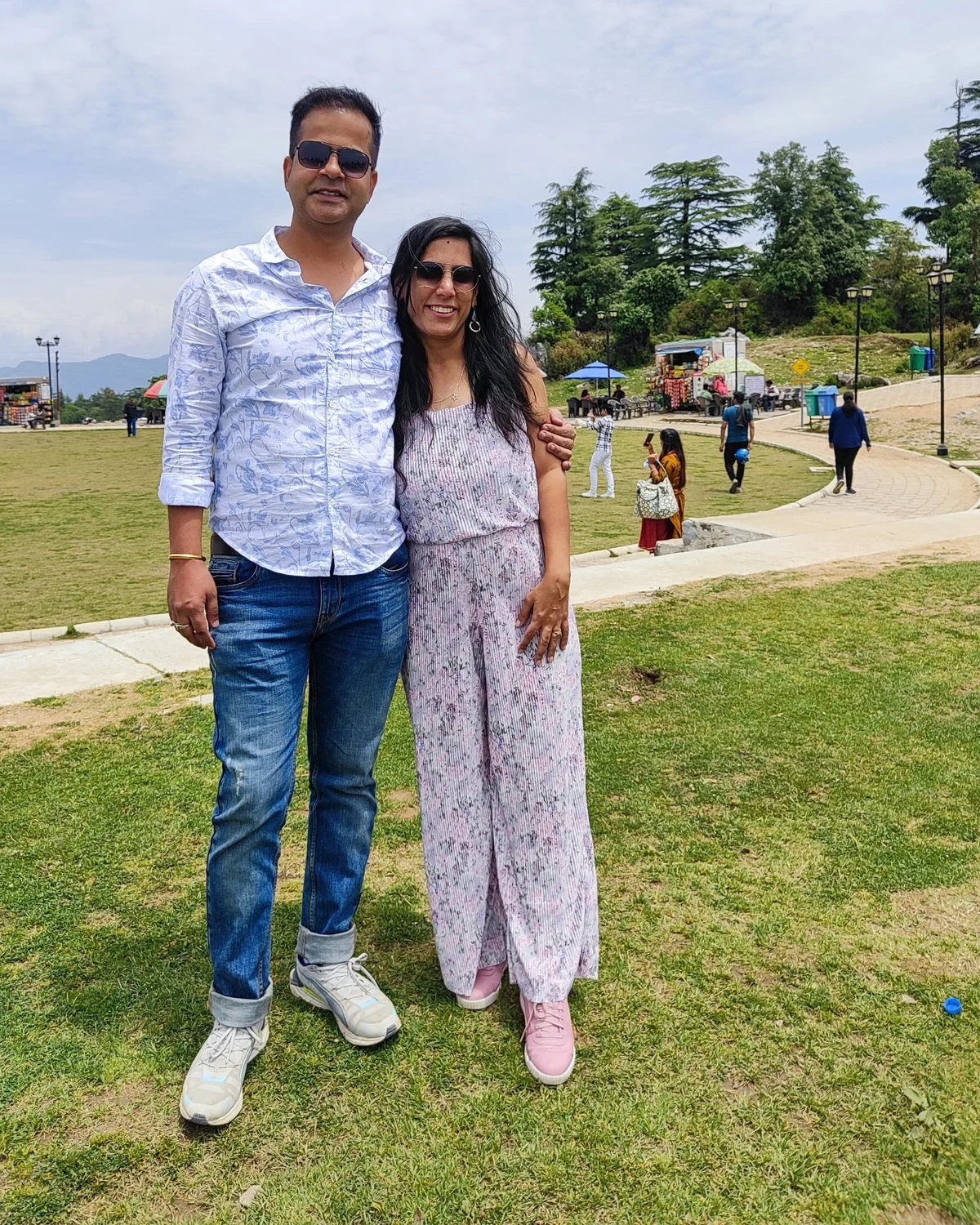 Poonam and Sunil standing together on a hilltop lawn ringed by deodar trees